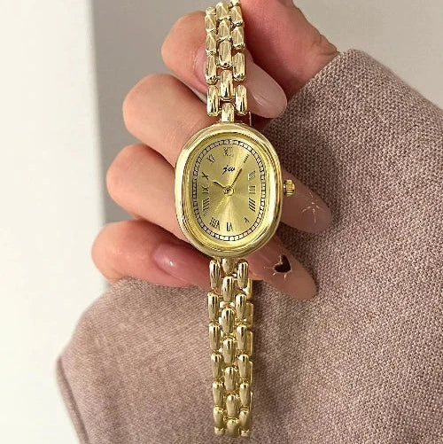 Gold watch held by a hand with beige nail polish on a neutral background